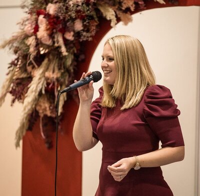 A woman with blonde hair is speaking into a microphone. She is wearing a maroon dress with puffed sleeves and is standing in front of a floral arrangement with red and pink tones.