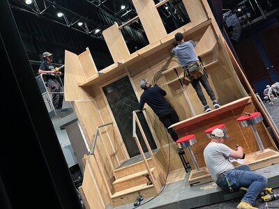A large wooden stage set under construction with framed platforms, railings, and stairs built by an experienced carpenter for a theater production.