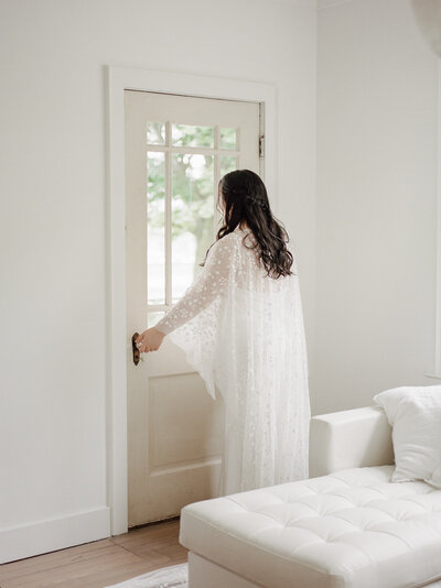 bride in patterned bridal cape opens door in clean white space, Michigan wedding venue Glass House Community