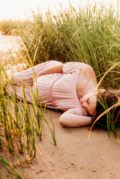 Expectant mother laying in the sand  amongst tall grasses.  She is cupping her belly bump and resting her head on her arm.  Photo taken by Philadelphia maternity photographer, Kristi