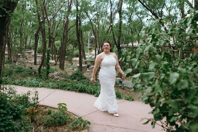 A bride walks on a paved path with greenery all around her.
