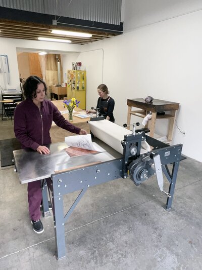 Two printmakers work together on a rolling press, transferring an etched design from a copper plate onto paper in a collaborative printing process.