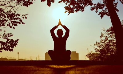 Silhouette of a person practicing yoga at sunrise in a seated pose with hands in prayer position, surrounded by trees and soft golden light—capturing the calm, spiritual essence of Enforma Tulum.