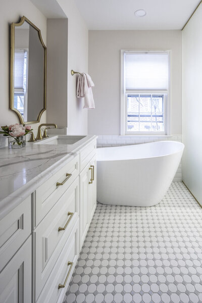 white octagonal and light gray square accent mosaic floor tile, pale pink walls, white slipper tub, white vanity with brass faucet and drawer pull hardware, brass mirror, pale pink bath towels, peony in vase on vanity, marble-look quartz countertop, frosted glass room divider