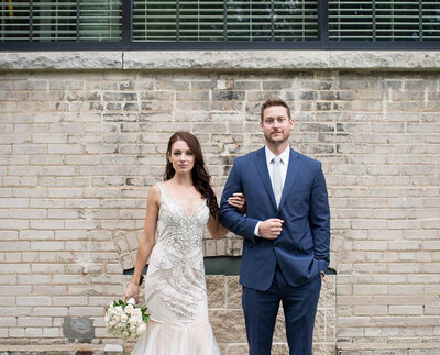 bride and groom standing against brick wall look seriously toward camera