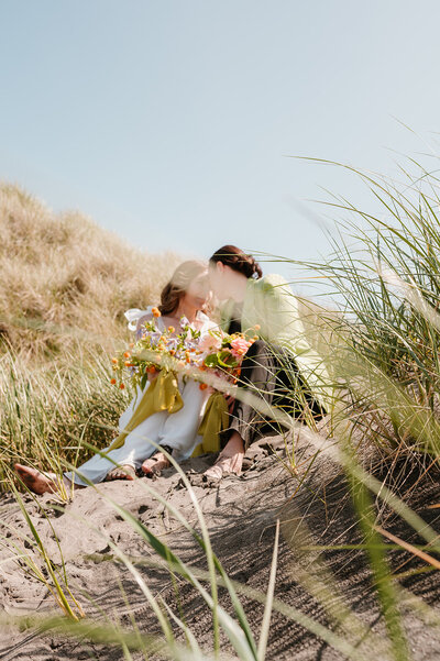 A candid wedding photo of a couple sitting together in the sand dunes on the Oregon Coast. They are two women holding their wedding bouquets and leaning in for a kiss. One woman is wearing a white wedding dress and the other woman is wearing a light green suit jacket with a black shirt and black pants.