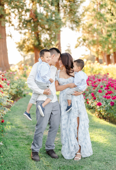 A family of four dressed in grays and greens stand in a field of roses as the mom and dad touch foreheads while holding their toddler sons photographed by Bay Area Family Photographer, Light Livin Photography.