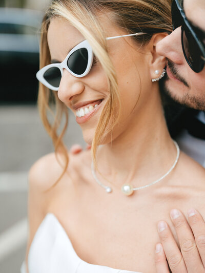 Stylish bride wearing retro white cat-eye sunglasses and pearl necklace smiles during romantic outdoor photoshoot.