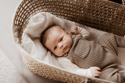 newborn baby boy in a knit over all out fit lays in a wicker Moses basket