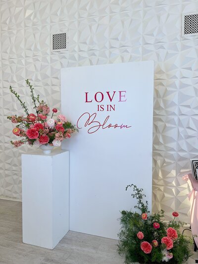 Modern event backdrop with the phrase ‘Love is in Bloom’ paired with pink floral arrangements, styled by Yours Truly, a wedding planner and designer in New Jersey.