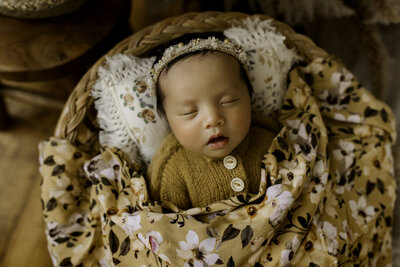 Sweet newborn baby girl in a yellow outfit and flower crown wrapped in a floral blanket.