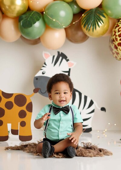 A joyful baby boy sits in a safari-themed setup with playful animal cutouts and balloon decorations. He wears a mint green shirt with a bow tie and suspenders, perfectly complementing the lively and fun atmosphere.
