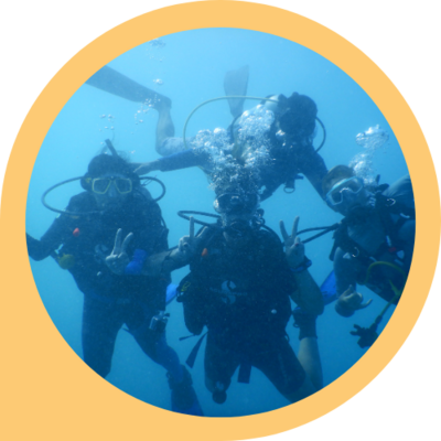 Four scuba divers pose for a photo underwater, dressed in wetsuits with masks and flippers. Bubbles rise from each of them as one makes a peace sign for the camera.