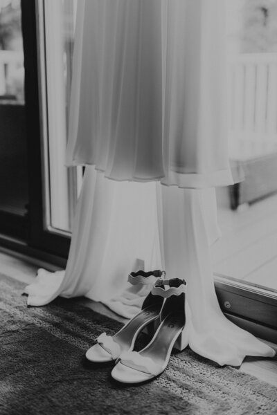 Black and white photo of wedding dress and wedding heels hanging in front of a sliding glass door.