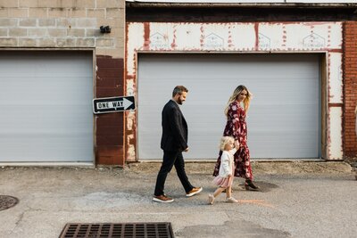 A family of three holding hands and walking past a garage with a "one way" sign, captured beautifully in a family photoshoot in Pittsburgh.