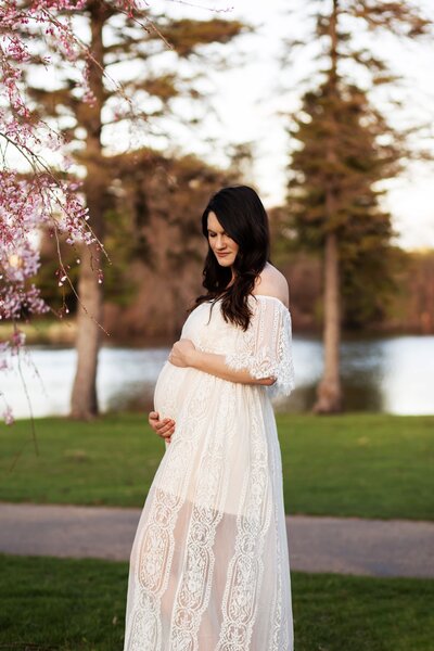 A pregnant woman wearing a lace dress looks down at her bump with a lake behind her.