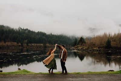 man and woman stand holding hands arms outstretched with lake and mountain behind them