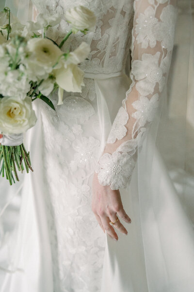 A romantic detail shot of a bride’s lace wedding gown and delicate gold engagement ring, captured at Dover Hall in Richmond, Virginia. The intricate floral appliqué on her sheer sleeve and the soft ivory tones of her bouquet evoke timeless elegance. Photographed by Caitlin Wilcox Photography, a luxury wedding photographer in Virginia specializing in film and digital imagery.