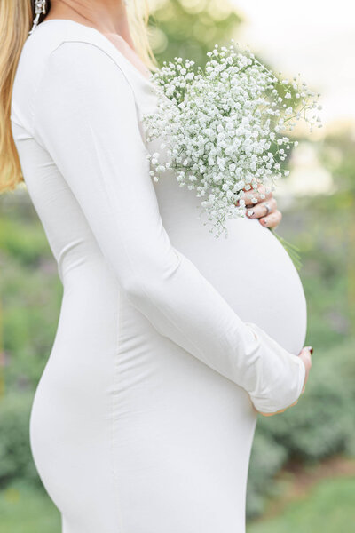 A mother holding a young toddler girl in a sun light rose garden as the toddler admires a soft pink rose she holds in her hand. 