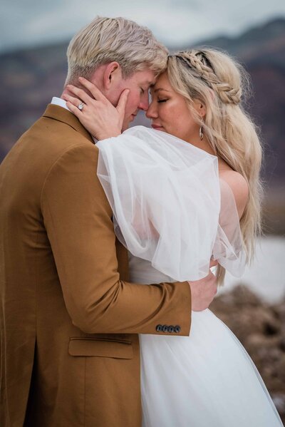 Adventure Elopement Package for Death Valley Elopements brought a bride and groom to a ceremony here so they are embracing in front of  a stark background with salt flats behind them.  YOu can see her ring as her hand rests on this neck and their foreheads touch