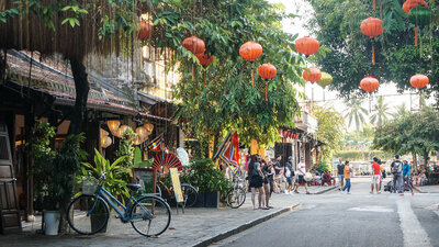A lively pedestrian street lined with shops, greenery, and bicycles, decorated with hanging red lanterns while people stroll and take photos.