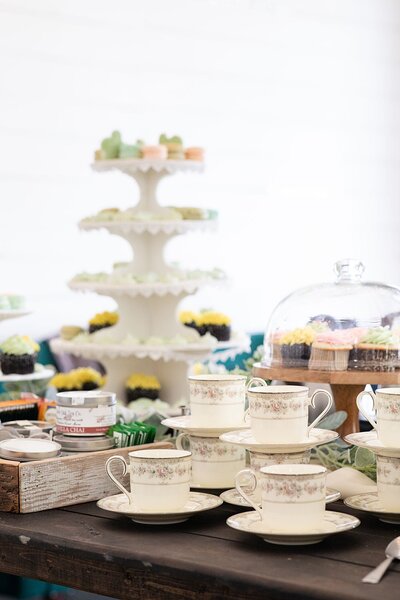 a stack of tea cups surrounded by different types of teas and a colorful display of cupcakes, macarons, and meringue kisses.