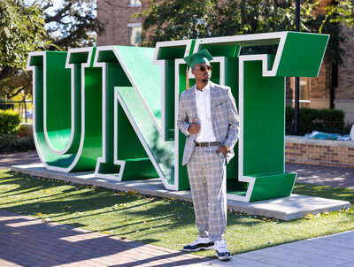 a college senior posing by a UNT sign while wearing a suit and his graduation cap