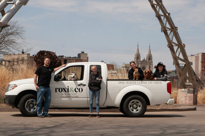 Roxi & Co team members posing with dogs in front of service truck at Sioux Falls Arc of Dreams with skyline in background.