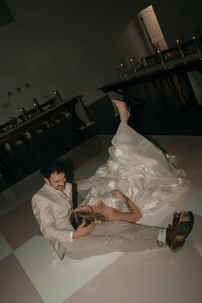 A couple in formal attire lies playfully on a checkered dance floor. The woman in a flowing dress has her legs playfully raised; the man, in a beige suit, is smiling warmly.