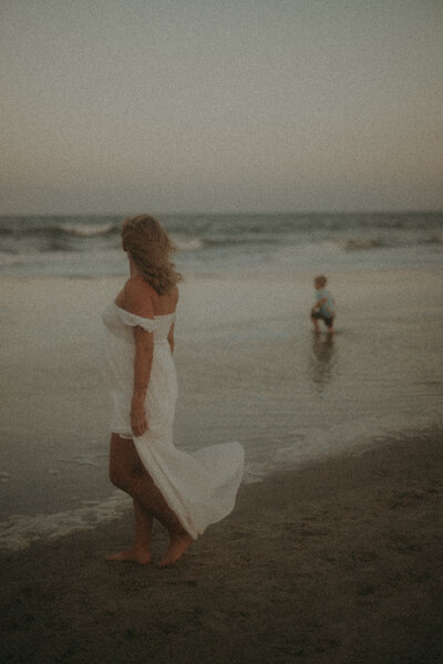A mother and child at the beach the mother is on the shore with her dress blowing in the wind facing away from the camera toward her son. The boy is further away walking into the ocean water