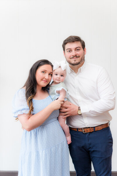 Mom in blue dress holding baby with Dad hugging around them.  Studio photo