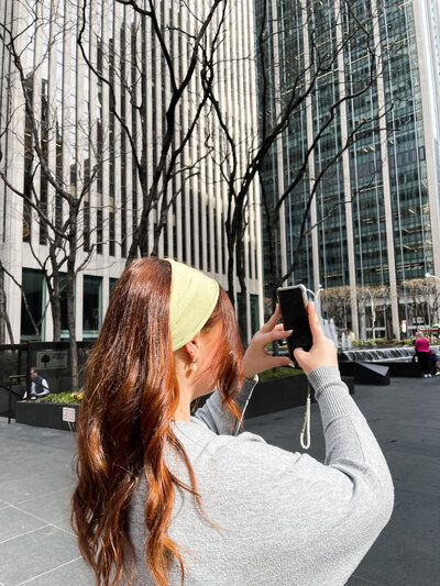 Woman with long red hair and a yellow headband taking a photo with a smartphone in a city plaza. Tall buildings and bare trees in the background.
