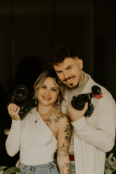 Laura et clément, souriants et enlacés, photographe et vidéaste de mariage, posent avec leurs appareils photo respectifs.