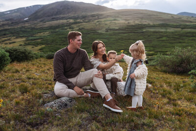Family with two young children sitting on the ground in the mountains. Little girl handing mom a flower.