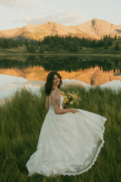 A bride in her wedding dress twirling with her bouquet on the edge of a glassy lake with mountain reflections.