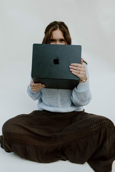 A girl sitting in front of a white backdrop holing her computer in front of her face. Showing her passion for showit website design.