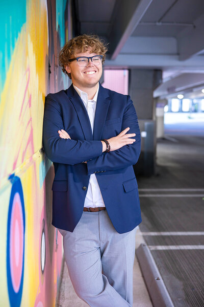 Caucasian senior boy wearing a navy blue blazer with a white shirt and gray dress pants, posing casually against a graffiti wall.