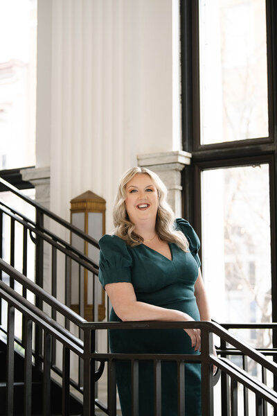 Career coach, Melissa Lawrence, leans on a staircase railing and smiles in a green dress