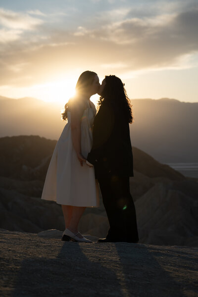 A newlywed queer couple kisses with a setting sun behind them