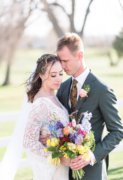 denver wedding photographer captures bride and groom embracing each other in a field on a farm for their outdoor summer wedding while bride wears a bohemian wedding dress and holds a bright floral bouquet