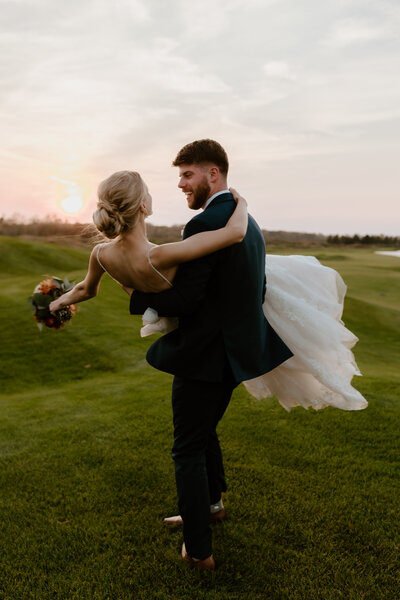 A groom in a dark suit joyfully carries a bride in a flowing white dress across a grassy field at sunset. The scene conveys happiness and romance.