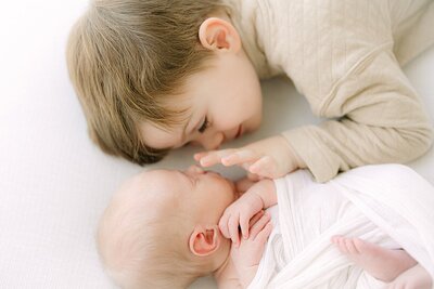 Toddler boy lays next to his newborn brother swaddled in white and gently touches his face during Indianapolis portrait photography session