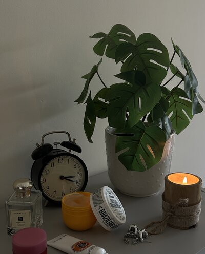 A cozy desk scene with a potted monstera plant, a lit candle, an alarm clock, and various skincare containers. The tone is calming and peaceful.