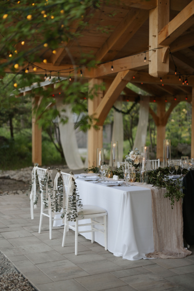 An elegantly set table under a wooden pavilion, decorated with white linens, greenery, and tall glass candle holders. String lights are draped across the wooden beams, and sheer white curtains hang in the background, creating a romantic atmosphere.
