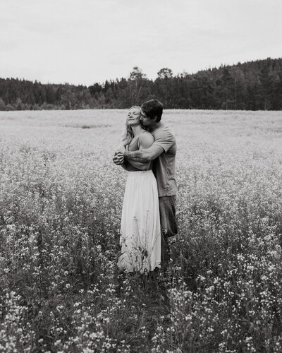 Man hugging his fiancé behind her back and she has her eyes closed at a rapeseed field in Finland