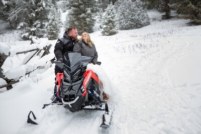 A bride sits on top of a snowmobile while her groom stands behind her smiling at her during their winter elopement in Montana.