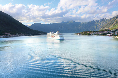 A large white cruise ship sailing through calm blue water surrounded by tall, green mountains under a partly cloudy sky.
