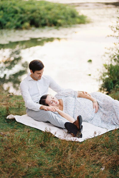 A pregnant mother is laying down on a blanket with her head resting on her husband's leg