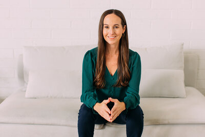 Kristie wearing a green blouse, sitting on a white couch, leaning forward and smiling