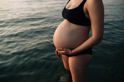 Pregnant woman in black bikini standing in water at beach.